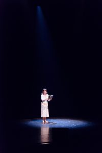 En kvinna i klänning står i strålkastarljus på en annars mörk scen.