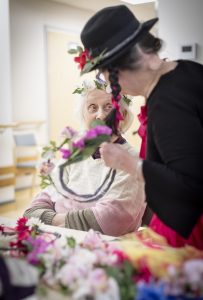 På bilden tittar en äldre dam på då en person utklädd till clown binder en blomsterkrans.