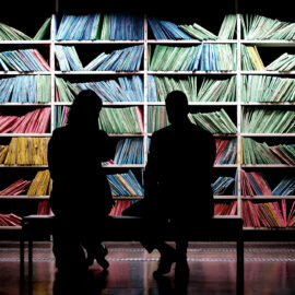 Två personer sitter på en bänk framför en bokhylla med böcker eller mappar sorterade enligt färg. De är vända mot bokhyllan. Personerna har ryggen mot kameran och man ser endast deras mörka silhuetter.