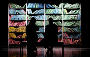 Två personer sitter på en bänk framför en bokhylla med böcker eller mappar sorterade enligt färg. De är vända mot bokhyllan. Personerna har ryggen mot kameran och man ser endast deras mörka silhuetter.