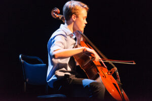En ung man som spelar cello på en nedsläckt scen.