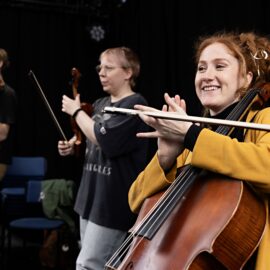 En cellist i gul tröja ler och klappar händerna med stråken mellan fingrarna. I bakgrunden knäpper håller en annan person sin violin som en gitarr och knäpper på den.