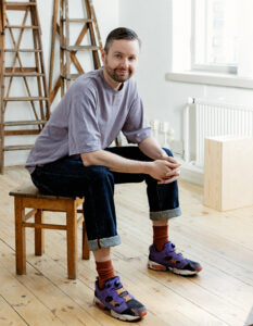 Regissören och koreografen Jakob Höglund sitter på en pall i en studio. Han har lila t-skjorta och uppvikta jeans. Han tittar in i kameran och småler.