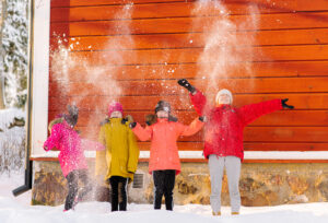 Fyra barn i färgglada kläder står på rad framför en röd trävägg och kastar snö upp luften. De blundar och ser glada ut.