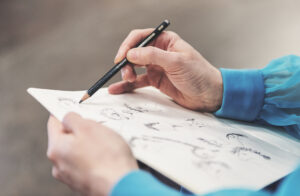En konstnär har ett skissblock i famnen och tecknar med blyertspenna på ett vitt papper. Endast konstnärens händer syns. Hen har en turkos långärmad blus.