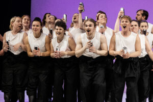 Fjorton personer iklädda vita linnen och svarta overaller står på en scen med en lila bakgrund. Personerna är riktade mot publiken. De håller i silverfärgade 0,33 liters burkar som de glatt skålar och skrålar med.