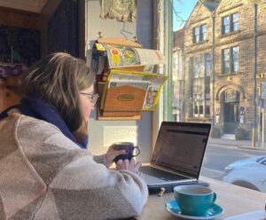 En person sitter vid sin dator. Hen har en vit-grå tröja och en kopp intill sin dator. Hen har brunt länger hår och vi ser henne snett bakifrån.