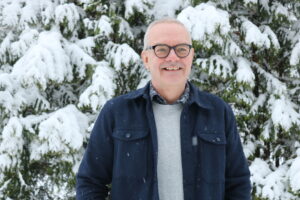 En bild föreställande en leende man i övre medelåldern. Fotot är taget utomhus och i bakgrunden syns snötäckta granar.