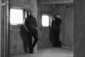 Ett svartvitt fotografi på två män i 30-års åldern som står i en avskalad och lite ruffig miljö som påminner om en lagerlokal.